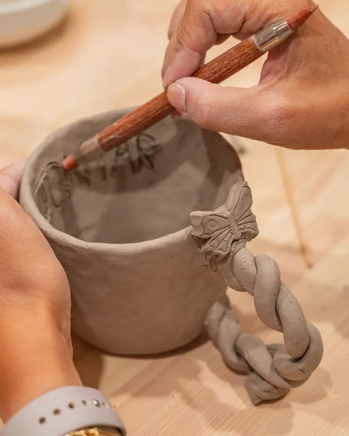 Hands carving a name into a hand-built cup with a braided handle