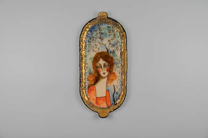 An elegant oval ceramic piece showcasing a female portrait with curly hair and a delicate tree motif in the background.